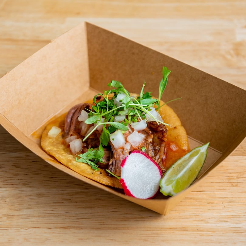 El Barrio Neighborhood Tacos' birria taco with cilantro, onions, radish, and lime in a cardboard tray.
