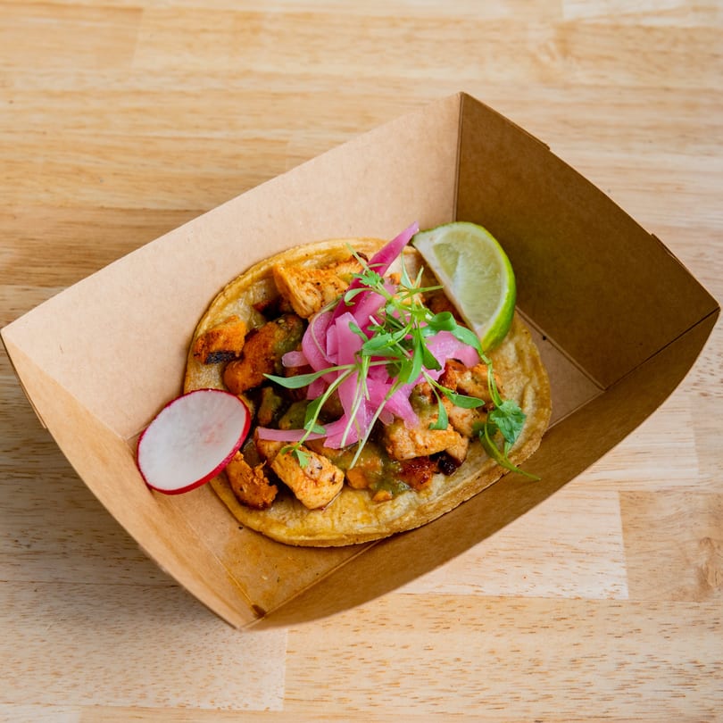 El Barrio Neighborhood Tacos' chicken taco with pickled onions, radish, and lime in a cardboard tray.